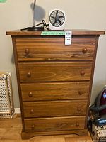 Wooden dresser with 5 drawers topped with a small fan; visible tags and measurements on dresser front