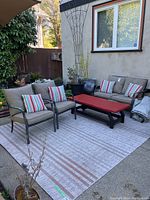 Full view of outdoor garden furniture set showing metal framed loveseat, two chairs with cushions and pillows, red wooden table, and outdoor rug (rug not included).