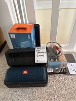 JBL Flip 5 Bluetooth speaker in carrying case with box and JBL Bluetooth headphones with charging cables and manuals on carpet.