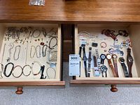 View of two open wooden drawers; left drawer contains various bracelets, necklaces, rings, and small items. Right drawer contains multiple wristwatches and some beaded necklaces.