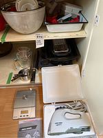 Photo showing shelf stocked with various baking and kitchen items including large metal mixing bowls, baking pans, glass mugs, muffin trays, and a Black & Decker handheld mixer with case.