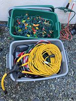 Two plastic bins filled with various electrical cords and string lights, showing coiled yellow extension cords and colorful Christmas lights with large bulb covers.