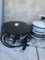 Full view of the fire pit and propane tank on concrete floor against wall, showing the stove knob, square base, hose, and strap.