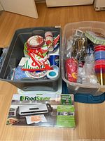 Two bins with assorted party supplies including paper plates, cups, napkins, and plastic cutlery, with FoodSaver vacuum sealer box in front