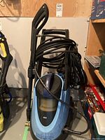 Front angled view of a blue electric pressure washer with attached black spray gun, coiled hose around the top handle, and wheels for maneuverability.