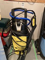 Full view of yellow and black Karcher electric pressure washer mounted on wheels with attached coiled hose and spray gun, showing front and left side of unit.
