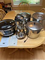 Photo showing all items on a dining table: Hamilton Beach slow cooker, Oster digital blender, and three large pots with lids, plus instruction manuals