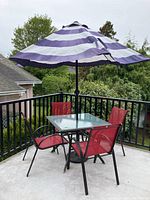 Patio set placed outdoors on concrete balcony with surrounding black metal railing; includes square glass-top table with umbrella installed, four red nylon mesh chairs.