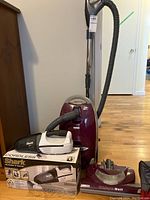 Kenmore canister vacuum with hose and metal wand, Whisper Belt floor head attachment, and Shark cordless handheld vacuum with its retail box on wooden floor.