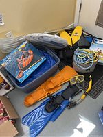 Photo of all water sports gear in a room corner showing double mattress float, inflatable boat box, yellow flippers, snorkeling goggles and snorkel, life jacket, and a bucket of ropes.