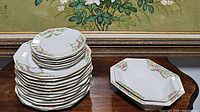 Stack of dinner and dessert plates plus two serving dishes on wooden surface in front of framed artwork.