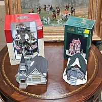 Two boxed village lighted houses with two unboxed ceramic buildings on table. Lemax Garden Shop in known packaging red box. General Store house in green box. Unboxed are Parish Church with steeple and Fishing Rental House with snow-covered roof and wreath.