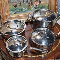 Set of four stainless steel Cuisinart pots with lids displayed on wooden surface with framed painting in background.