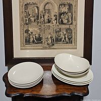 Stack of 5 cream-colored dinner plates and stack of 3 oval serving platters and 3 oval serving bowls on a wooden table with framed artwork behind.