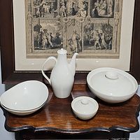 Five cream-colored china pieces arranged on a wooden table against framed artwork: one tall pitcher, two covered serving dishes, and two open bowls.