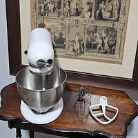 Front and side view of white KitchenAid mixer with stainless steel mixing bowl and two attachments on tabletop.