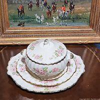 Covered vegetable bowl with lid and three matching serving platters arranged on a surface with a framed painting in the background. The china has a white porcelain base with pink rose floral designs and 22k gold accents on edges and handles.