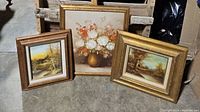 Three framed paintings standing on floor: two landscape cabin scenes and one floral still life.