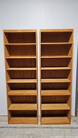 Pair of tall wooden bookcases with 6 shelves each, shown side by side against a plain wall, highlighting their height and shelf count.