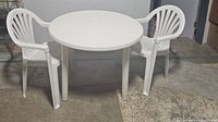 White round plastic patio table and two white Rubbermaid plastic chairs arranged on concrete floor indoors.