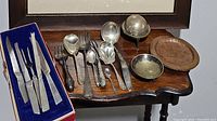 Overall image of silver and copper serving pieces on wooden table including knives, forks, spoons, copper plates, butter dish and decorative brass bowl.