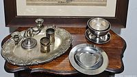 Silver serving ware displayed on wooden table, includes platter, candlestick holder, cylindrical dishes, tea/glass holder with saucer, and two round dishes.