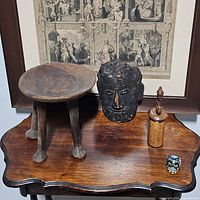 Overview of all four items on a wood surface in front of a framed print: stool, tribal mask, wooden container, sugar skull
