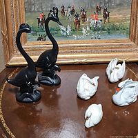 Photo showing all six swan figurines together, including two tall black mid century swans and four smaller white swan figurines, placed on a wood surface with a framed painting in the background.