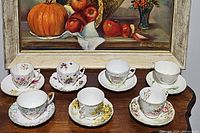 Photo showing all 7 teacups with matching saucers arranged on a wooden surface in front of a framed artwork.