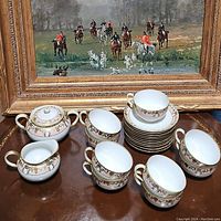 Photo of Nippon china set showing 5 teacups, sugar container, cream pitcher and stacked saucers arranged on a tabletop in front of a framed painting.