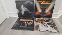 Four vinyl record covers arranged on floor showing front artwork including John Lennon & Yoko Ono Double Fantasy, ZZ Top Eliminator, Elton John Don't Shoot Me I'm Only the Piano Player, and Elton John Greatest Hits.
