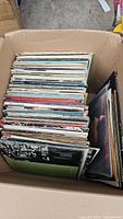 Top view of a cardboard box filled with numerous vinyl records with visible cover edges in various conditions.