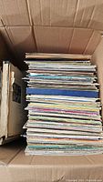 Stack of assorted vinyl records inside a cardboard box showing various album edge colors and sleeve conditions.
