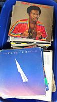 Top view inside large blue bin showing records with Arrow album cover featuring a man in a red track jacket and multicolor stripes.