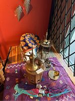 Overall view of purple Berber rug on floor with leather pouf, decorative lantern, small brass bowl, textile tassels and wooden side table with brass ewer and cups.