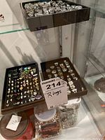 Two trays of assorted rings displayed in glass cabinet, top tray with silver-tone chunky rings, bottom tray with mixed metal fashion rings and colored stones