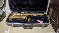 Alto saxophone body resting in open hard case lined with blue velvet alongside neck piece