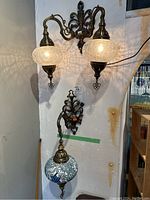 Full view of the two Turkish wall sconces mounted on wall. One sconce has two clear textured glass globes, the other has a blue mosaic globe.