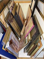 Photo shows multiple framed prints and canvas art pieces stacked together, with some pieces large and others smaller, all dusty from storage.