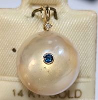 Front angled view of round cream-white freshwater pearl pendant with a blue gemstone inset and diamond above the bail, mounted on 14KT gold.