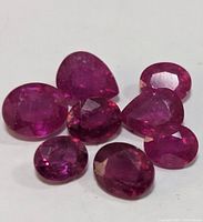 Seven loose genuine rubies arranged on white background, showing various shapes and sizes with faceted cuts and rich red color.