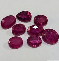 Eight loose faceted ruby gemstones of various oval, pear and round cuts displayed on white surface.