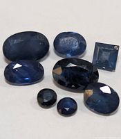 Seven faceted genuine sapphire gemstones in various shapes and sizes displayed on a white background.