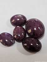 Five polished oval-shaped star rubies grouped together on a plain surface, showing their deep purplish-red color and reflective luster.