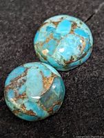 Two genuine turquoise cabochon gemstones with visible brown matrix pattern on a black textured background.