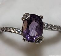 Close-up front view of sterling silver ring with oval faceted purple amethyst and three decorative prongs with small clear stones, plus textured silver band.
