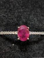 Close-up top view of the sterling silver ring with an oval ruby center stone and clear stones on the band.