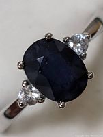 Close-up front view of dark blue oval sapphire with two clear round side stones, set in polished sterling silver band.