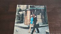 Front cover of John Lennon 45rpm single "Watching The Wheels," showing John Lennon and Yoko Ono walking on a street in a color photograph.