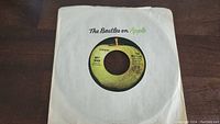Photo of a 45rpm vinyl record by The Beatles featuring the yellow/green Apple label, in a paper sleeve marked 'The Beatles on Apple' with some wear.